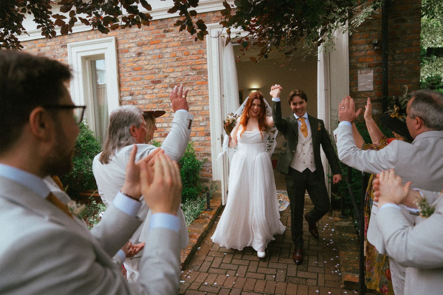 Bride and groom celebrated by cheering wedding guests outside brick venue.