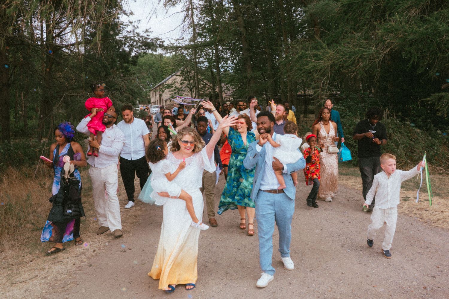 Wedding party celebrating outdoors on a dirt path, dancing joyfully among trees with guests in colorful attire.