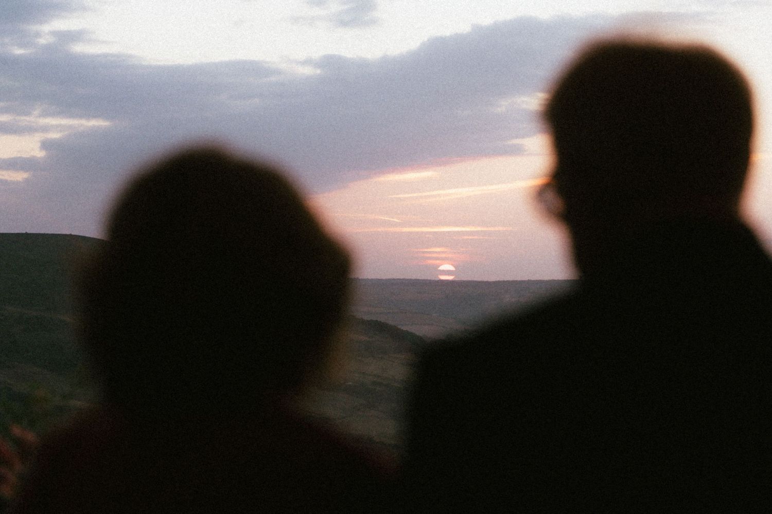 Two silhouetted people stand on a hillside watching a glowing orange sunset sink toward the horizon over the sea under soft clouds.
