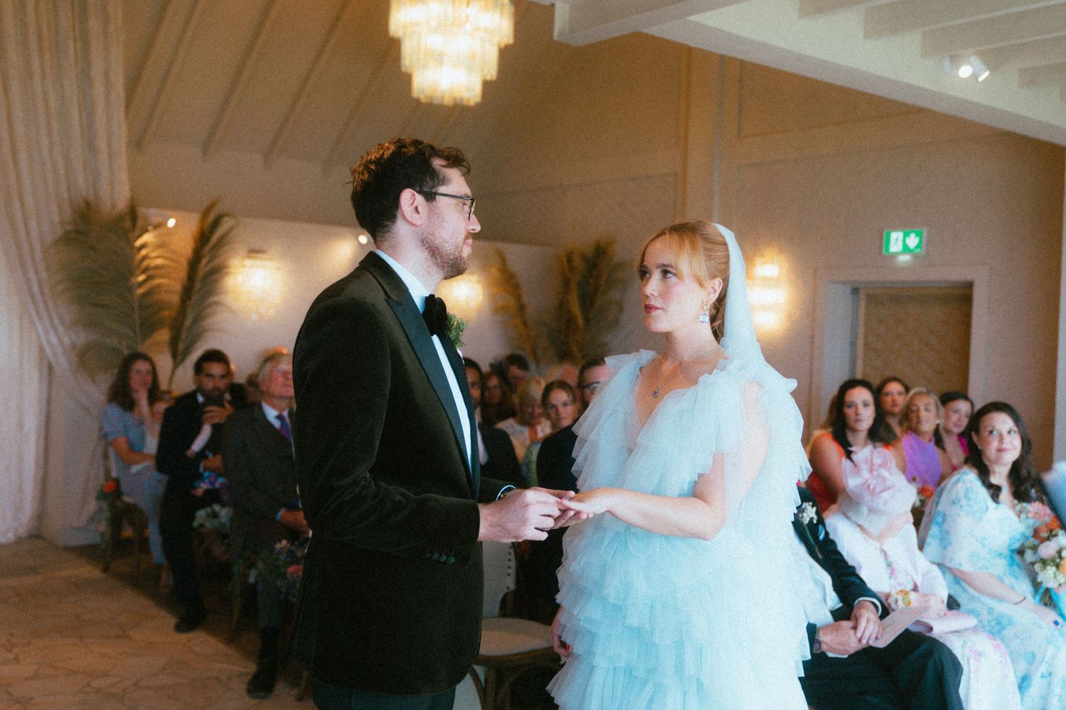 Groom in a dark velvet jacket and bow tie holds hands with a bride in a pale blue tulle gown as wedding guests watch during the ceremony.