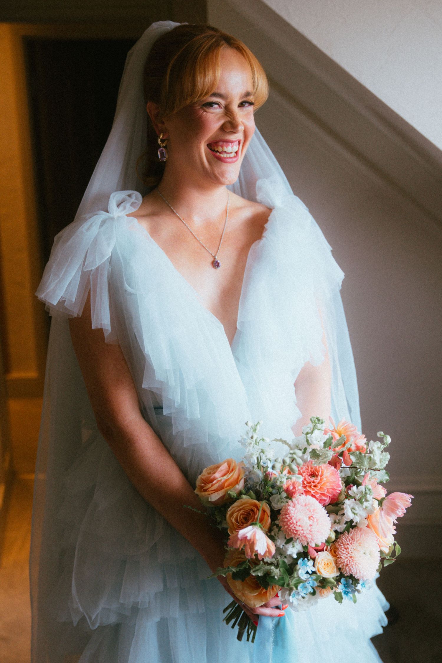 Smiling bride in a light blue tulle wedding dress and veil with bow details holding a peach and pink floral bouquet by a window.