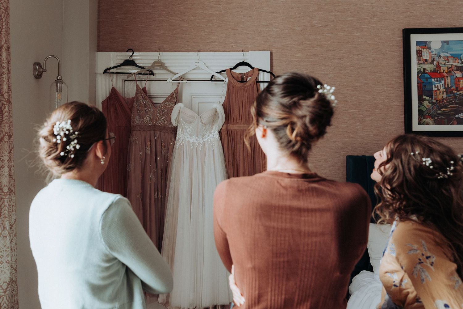 Bridesmaids admiring wedding dress and dusty rose bridesmaid gowns hanging in hotel room.