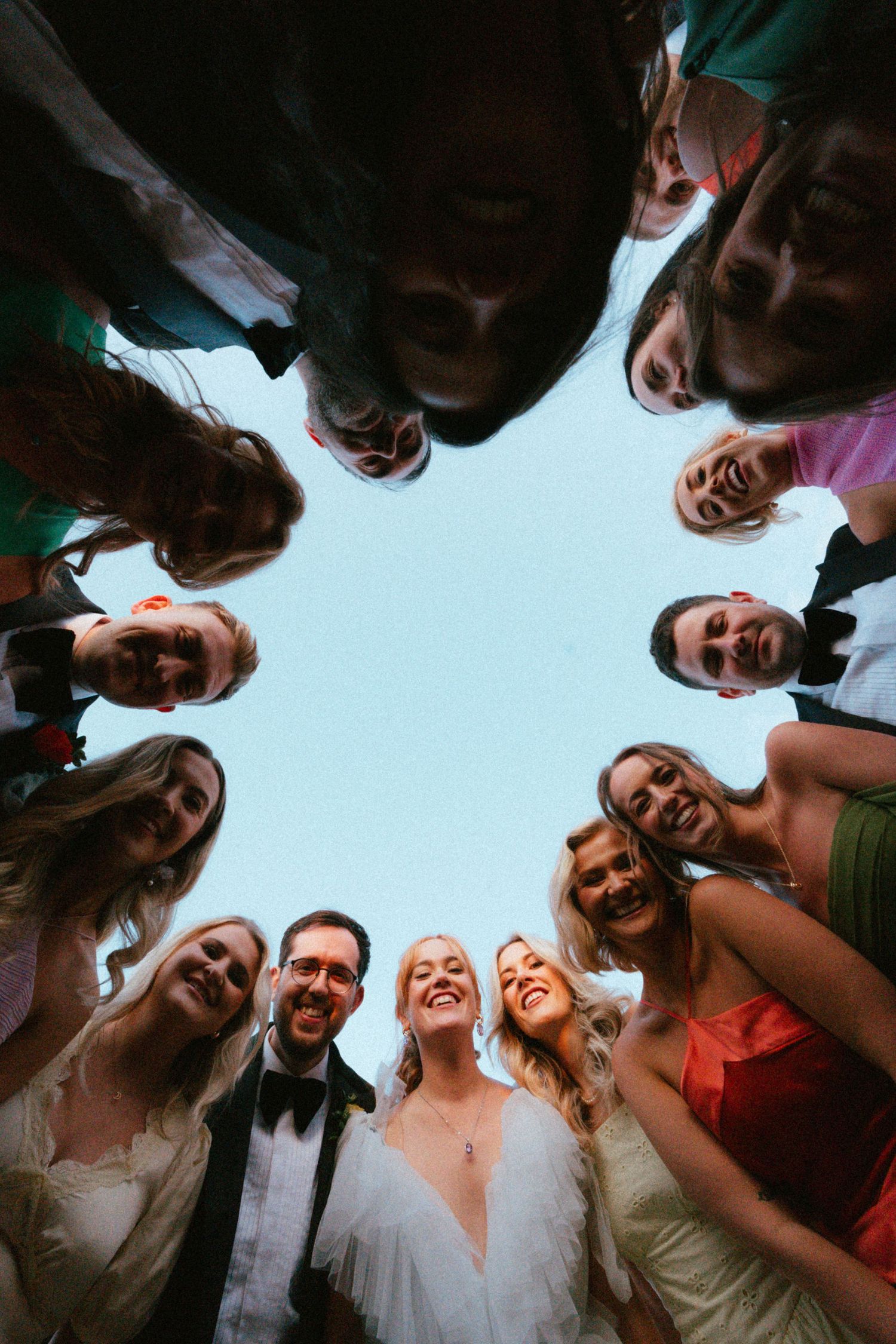 Low-angle view of smiling wedding guests in formal attire huddled in a circle, looking down at the camera against a pale sky.