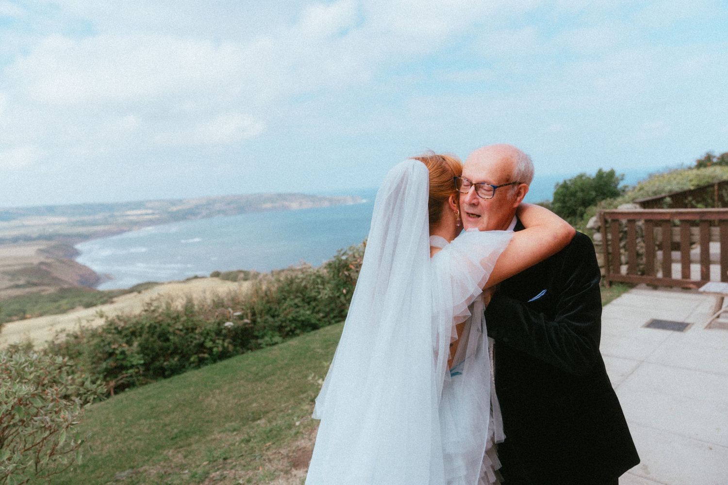 Bride in a white wedding dress and long veil hugs an older man in glasses and dark suit on a grassy clifftop overlooking the sea.