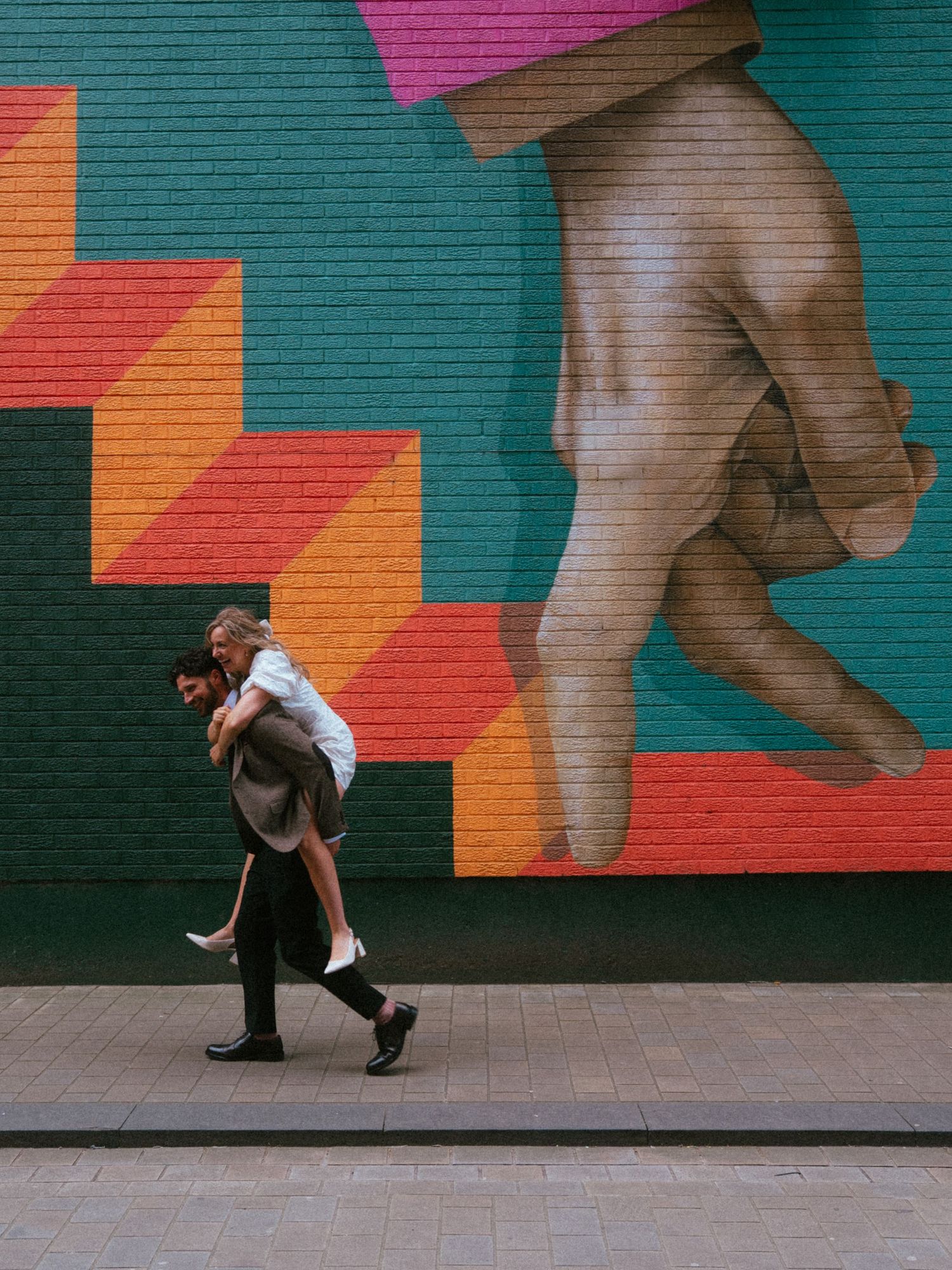 A couple playfully piggybacks in front of a vibrant colorful mural featuring geometric shapes and a large hand on a brick wall.