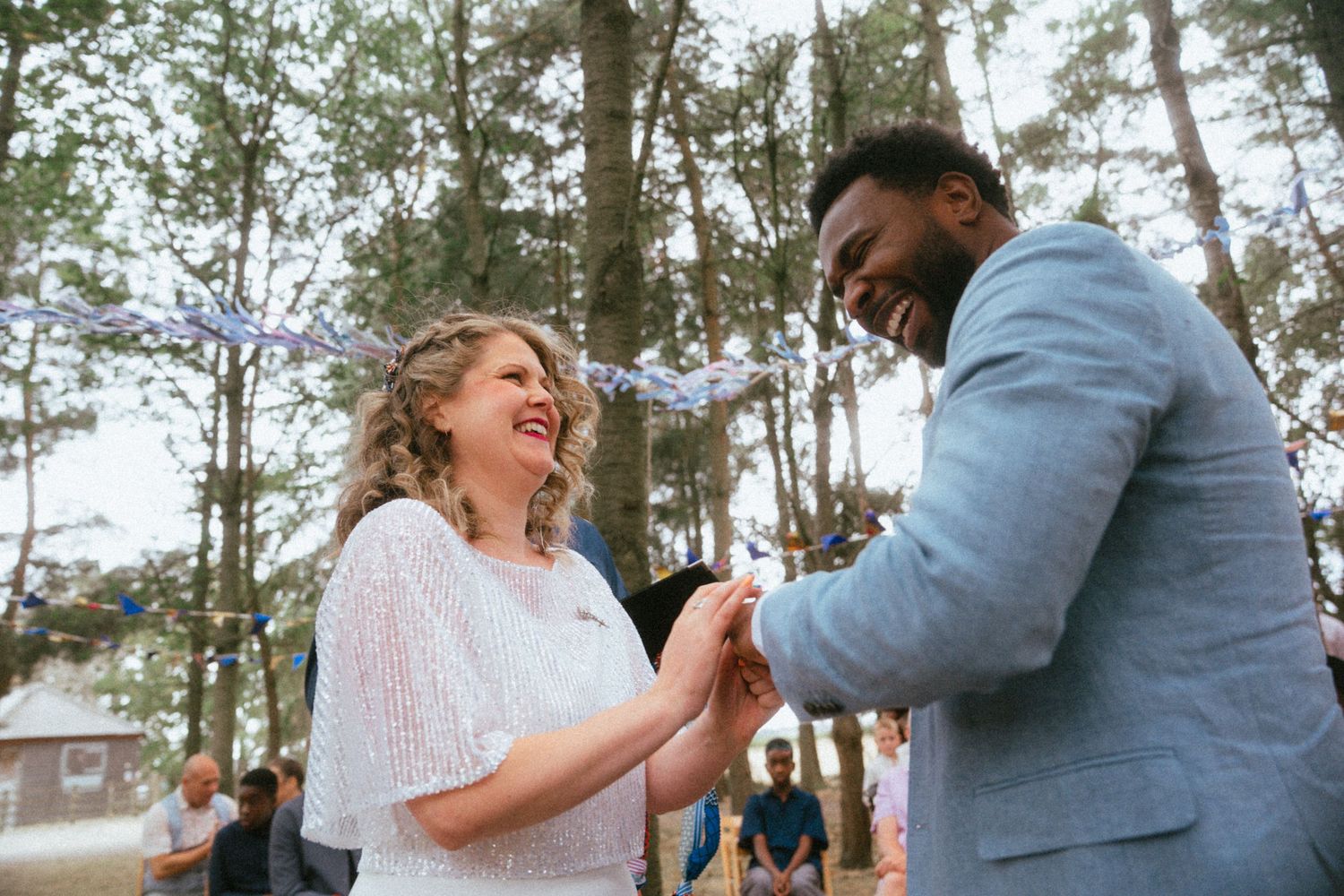 Couple laughing and holding hands during outdoor wedding ceremony in forest with colorful bunting.