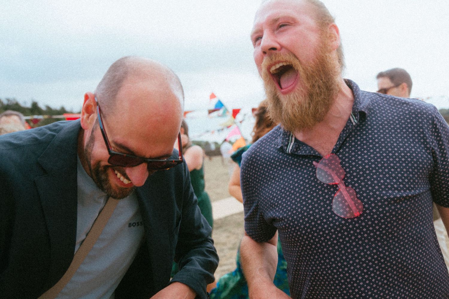 Two bearded men laughing outdoors at a festival, one bald in a suit, the other in a blue shirt with sunglasses.