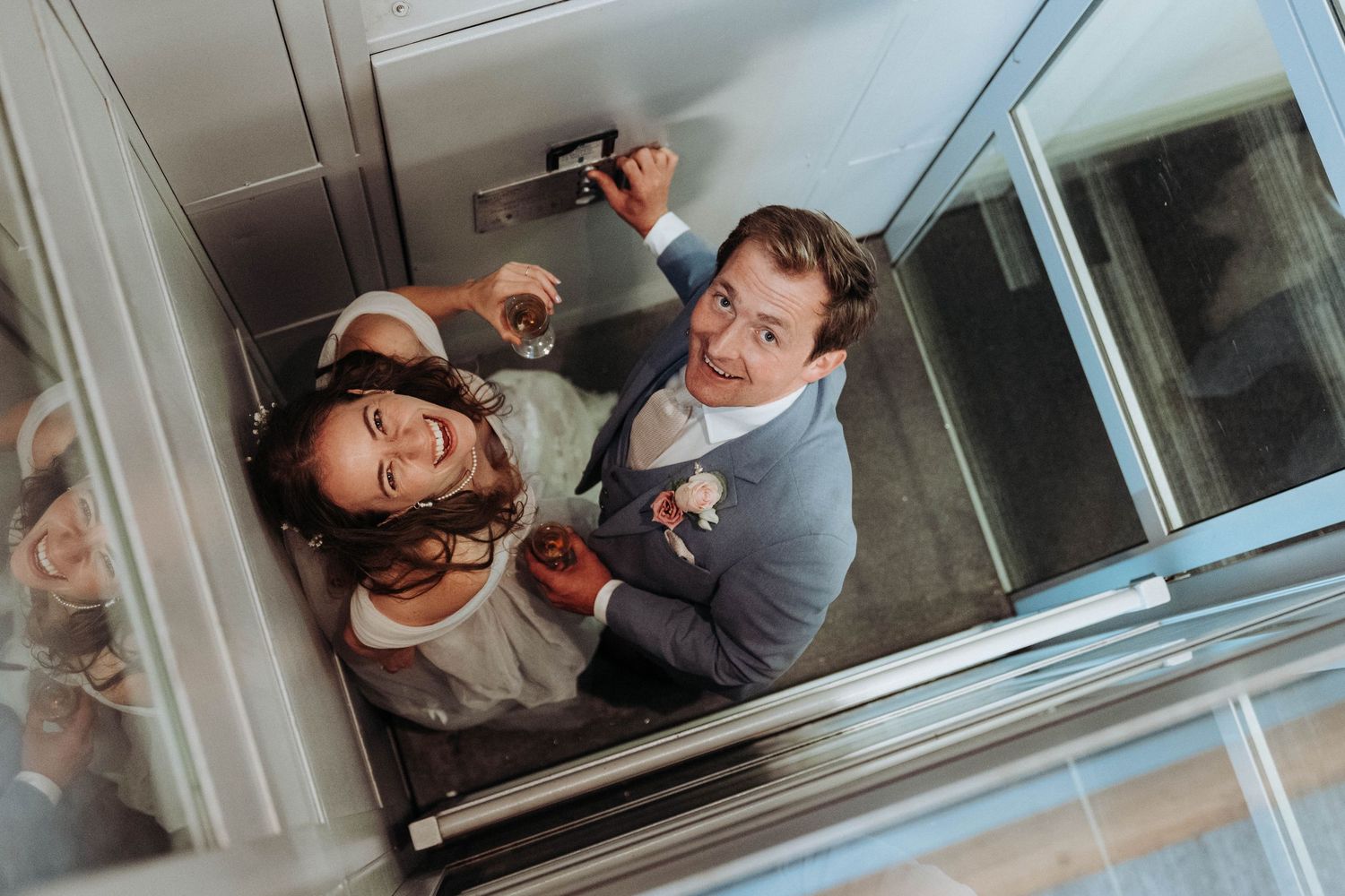 Bride and groom smile up at camera from inside a glass elevator at wedding venue.