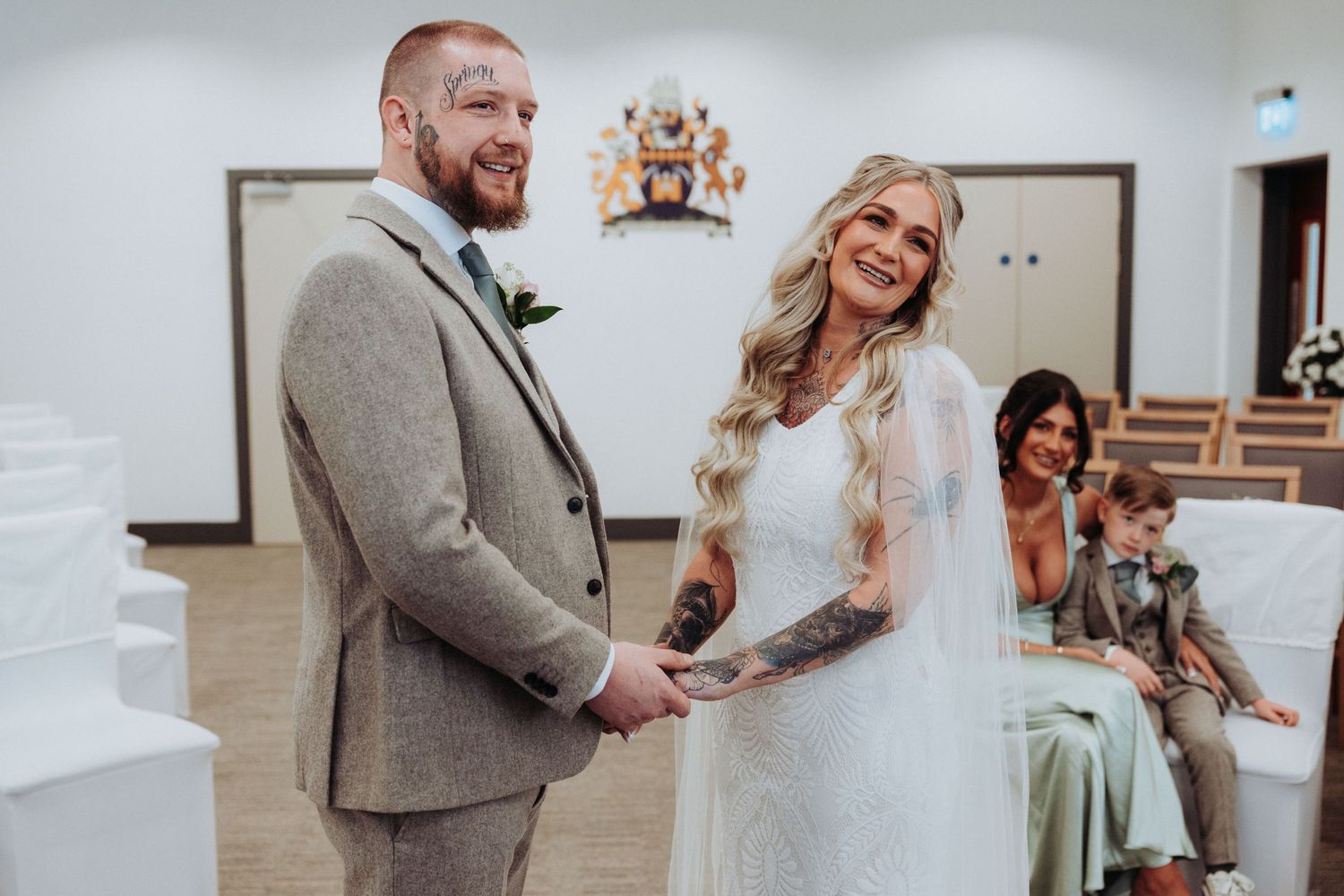 Smiling bride and tattooed groom holding hands at civil wedding ceremony in bright modern register office.