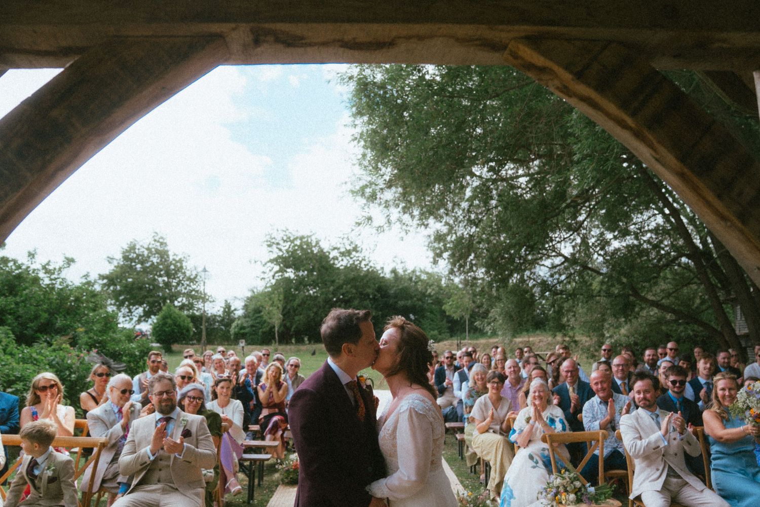 Bride and groom share first kiss under rustic wooden barn roof with seated wedding guests outdoors.