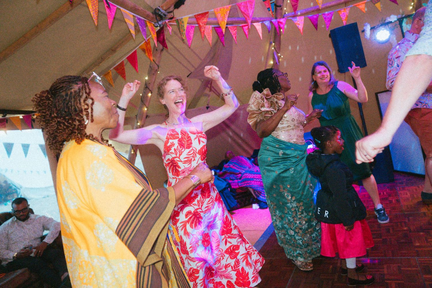 Joyful wedding guests in colorful dresses dance together under bunting lights at a lively reception celebration.