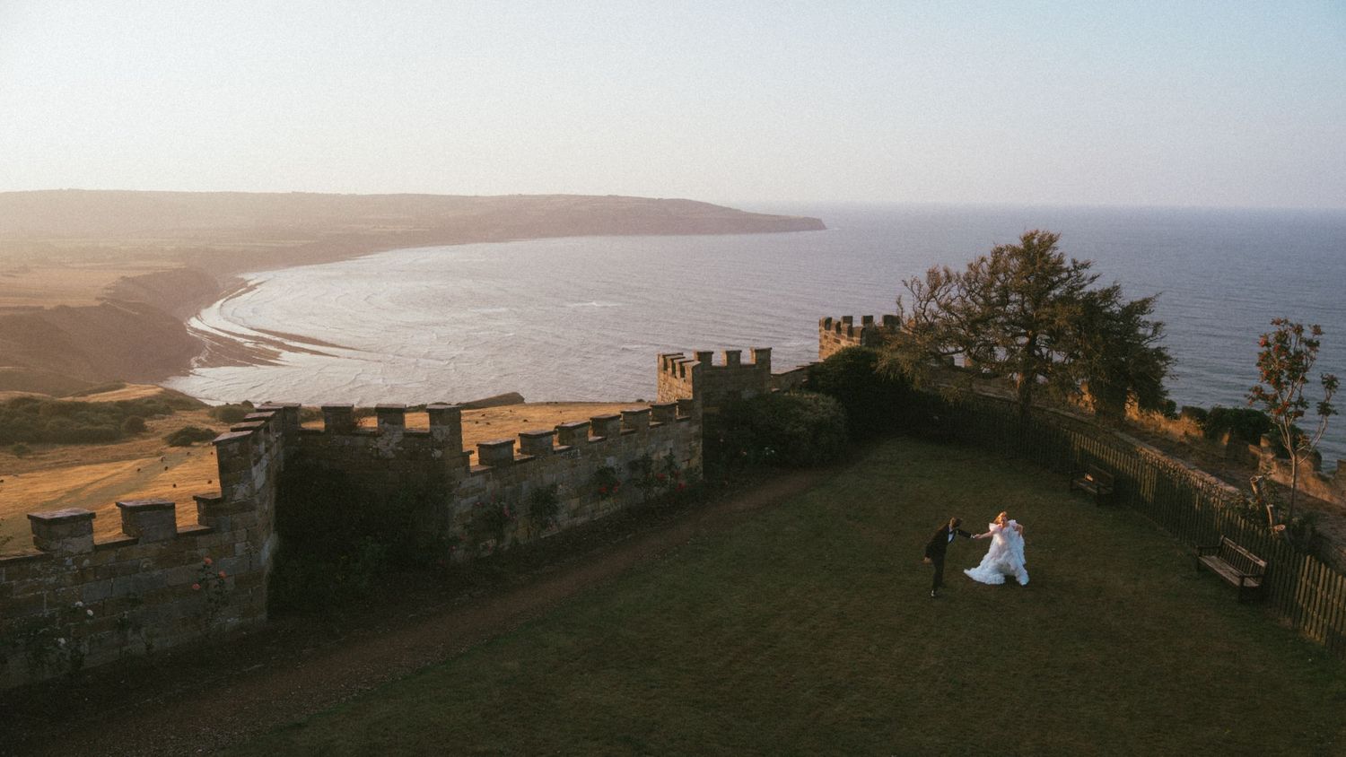 Aerial view of a bride and groom on a grassy lawn beside a castle wall overlooking the sea at sunset.