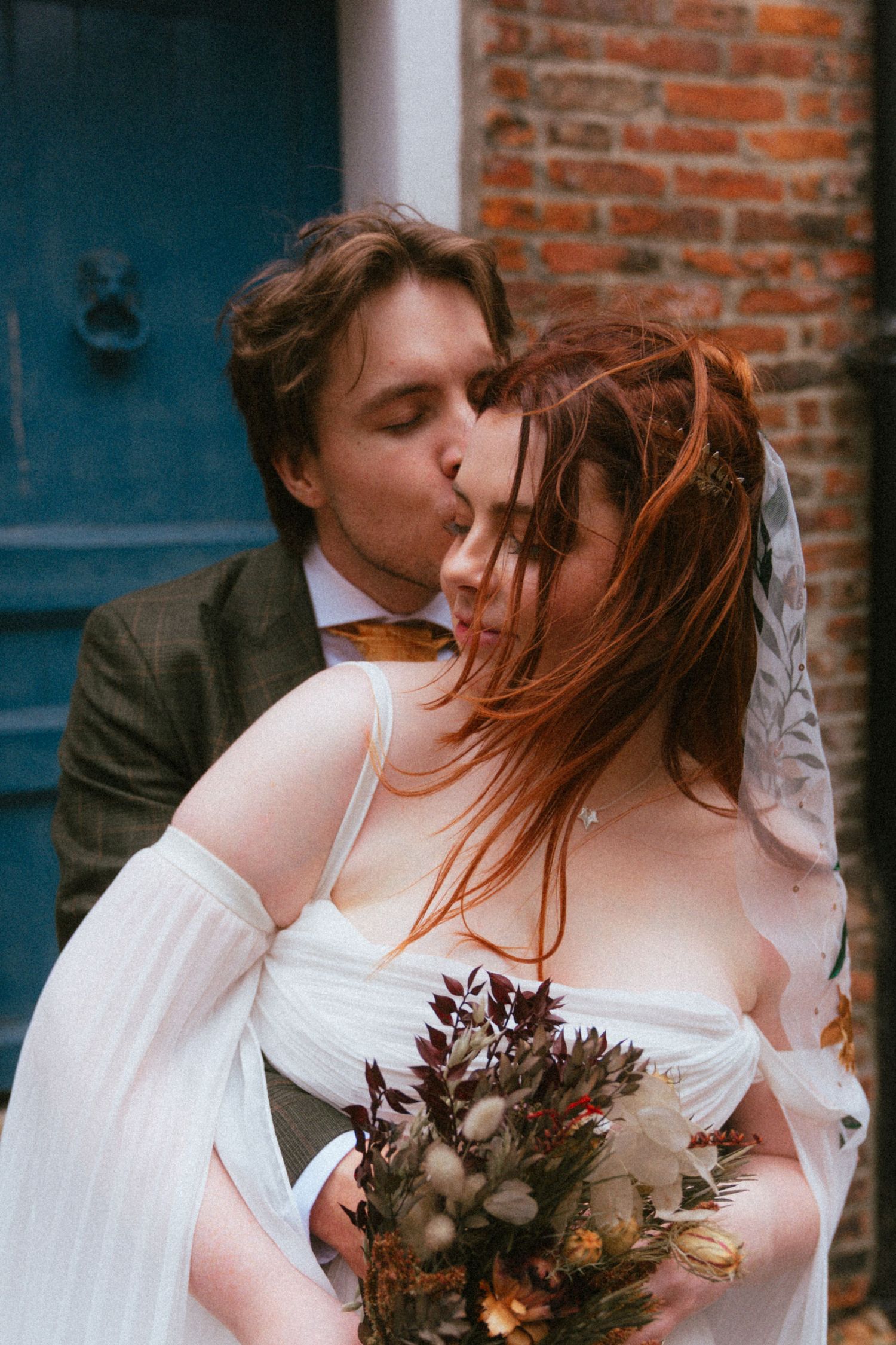 Bride and groom share romantic kiss outside blue door holding autumn bouquet.