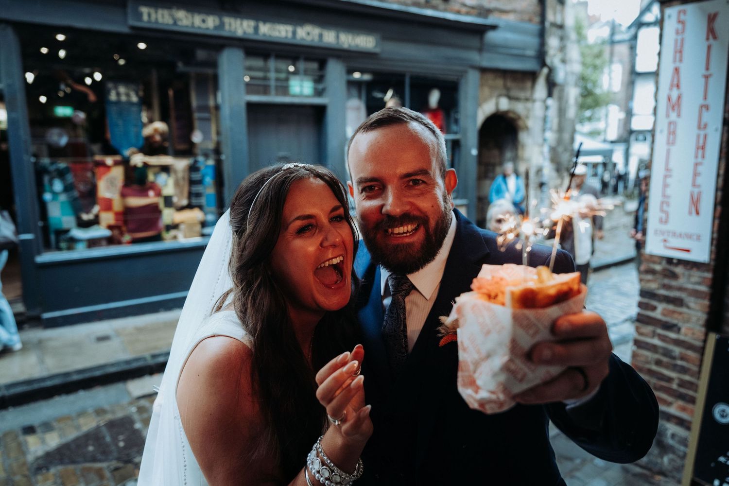 Excited newlywed couple holding sparkler dessert treat on cobblestone street after wedding ceremony.