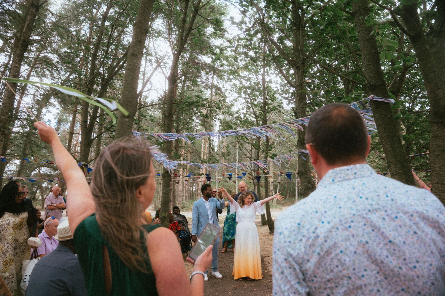 Wedding guests cheering and waving as couple walks through a tree-lined outdoor ceremony space with festive bunting.