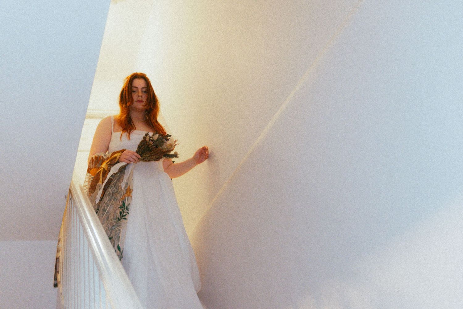 Bride in a flowing white gown holding a bouquet while descending a staircase on her wedding day.