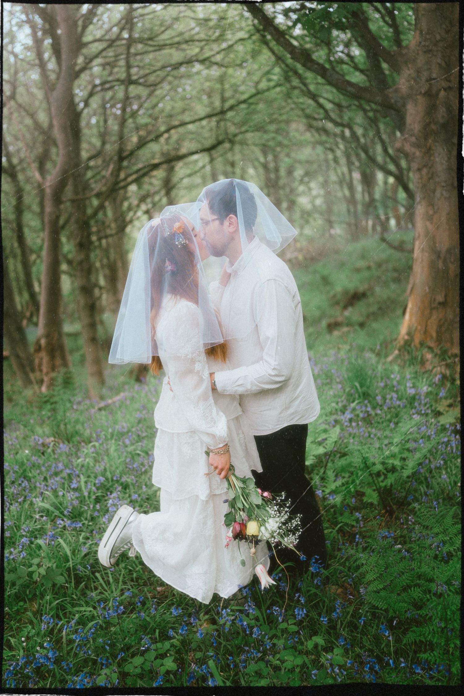 Bride and groom kissing under a veil in a bluebell woodland, bride holding a bouquet in a white dress.