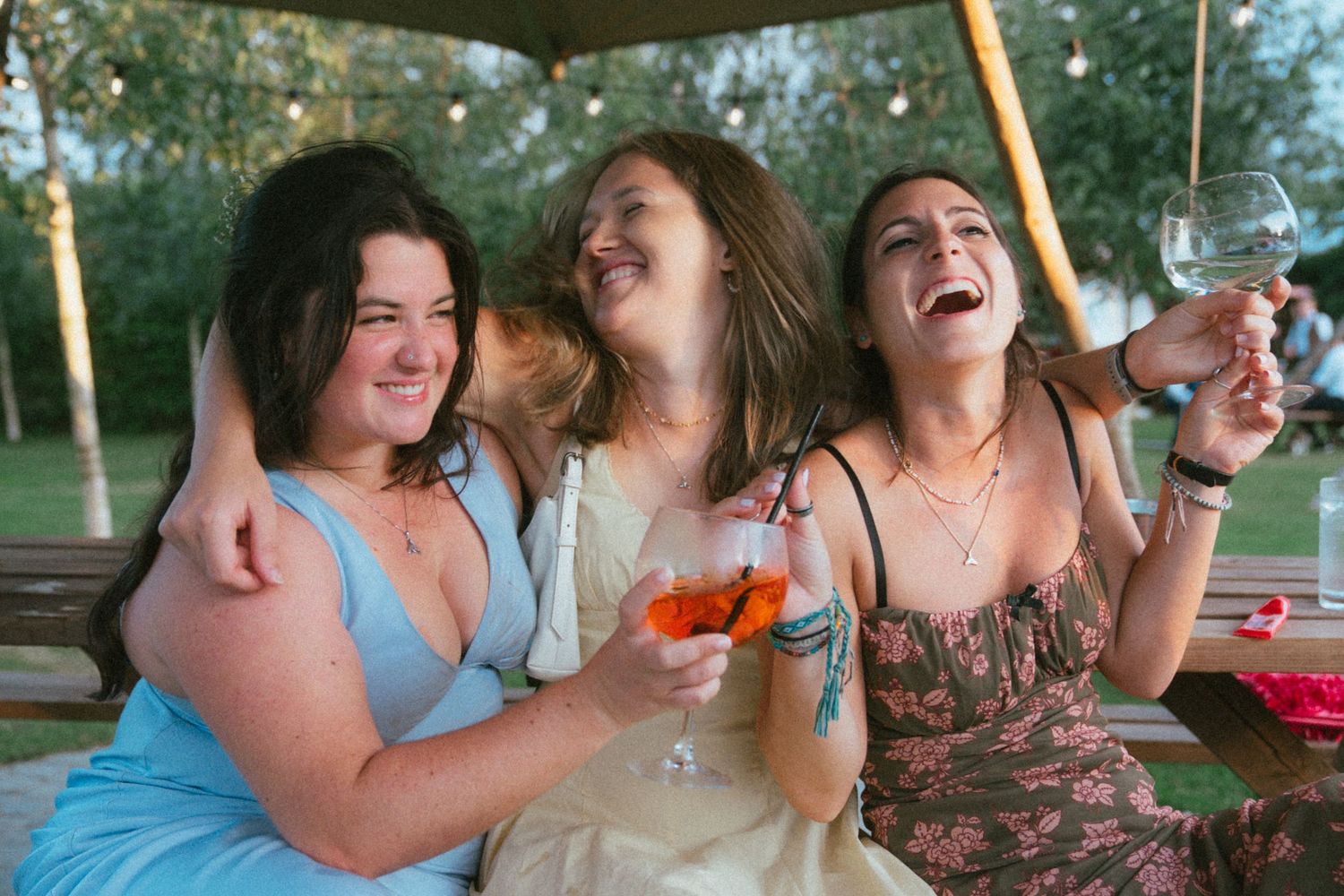 Three women laugh joyfully together holding drinks at an outdoor wedding celebration.