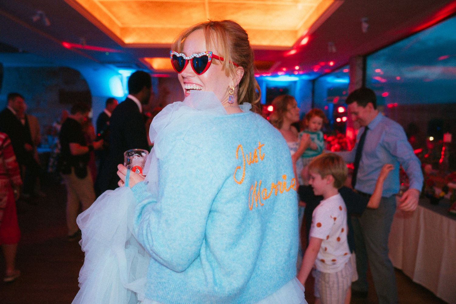 Smiling bride in heart-shaped sunglasses and a light blue jacket dances with guests at an indoor wedding reception.