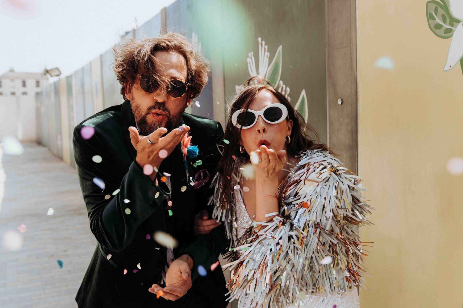Couple blowing confetti wearing festive outfits and sunglasses during a celebration outdoors.