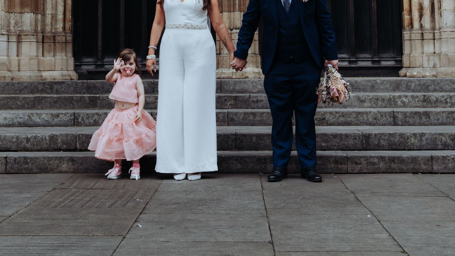 Bride in white jumpsuit and groom in navy suit hold hands with flower girl on cathedral steps.