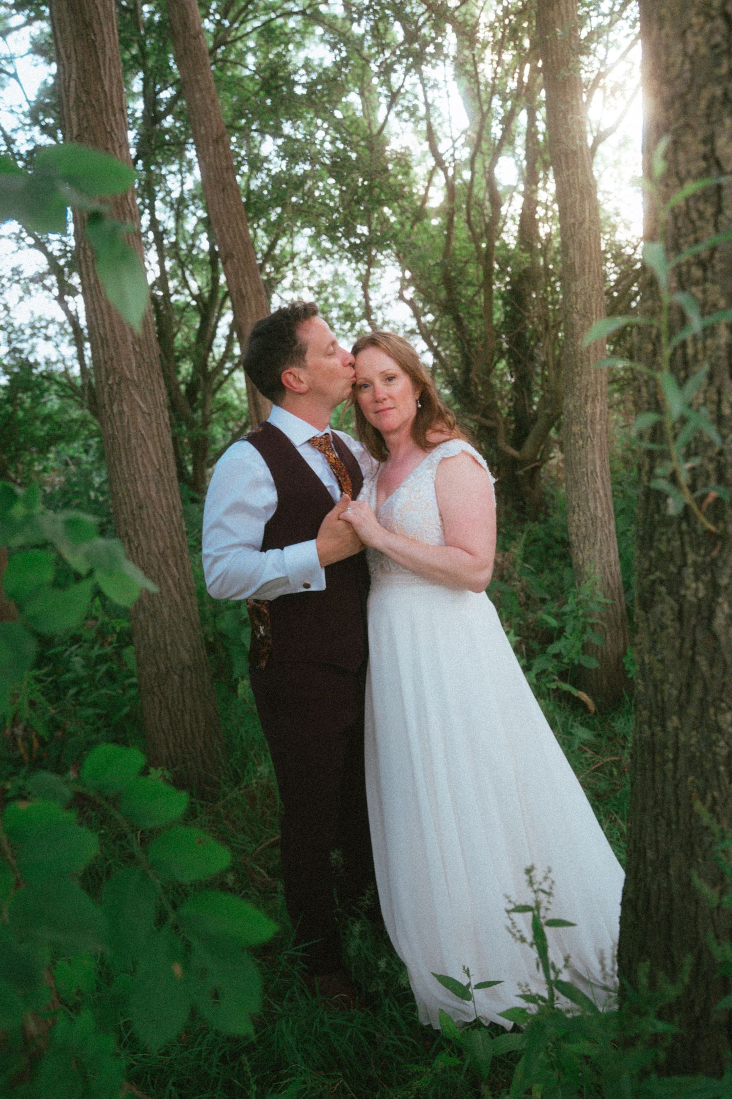 A bride and groom share a romantic moment among lush green trees in a sun-dappled forest setting.