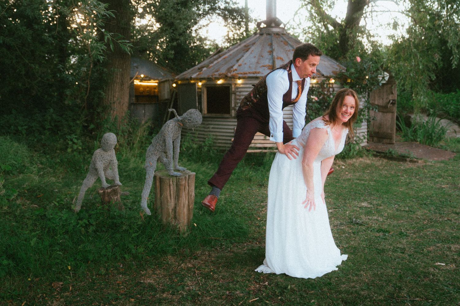 Bride and groom pose playfully outdoors near rustic gazebo with whimsical garden sculptures at twilight wedding celebration.