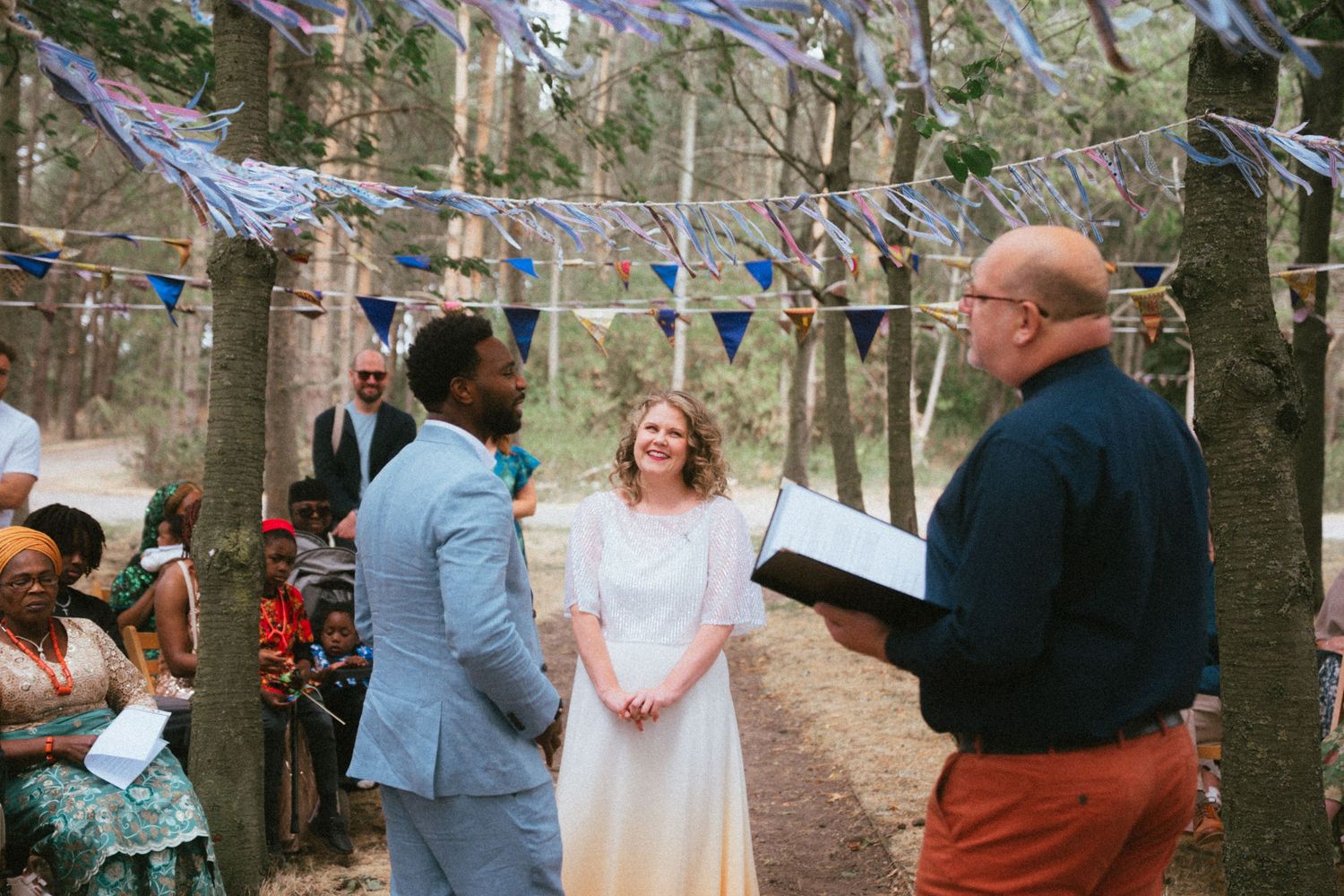 Couple exchanges vows outdoors in forest wedding ceremony with colorful bunting decorations and officiant reading from book.