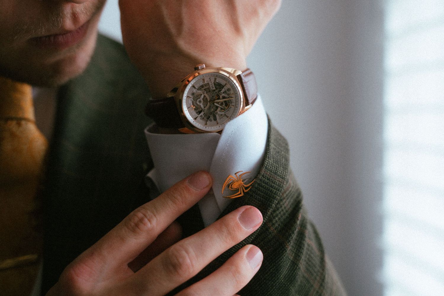Man in green suit adjusting brown leather watch with rose gold case and exposed mechanical movement.