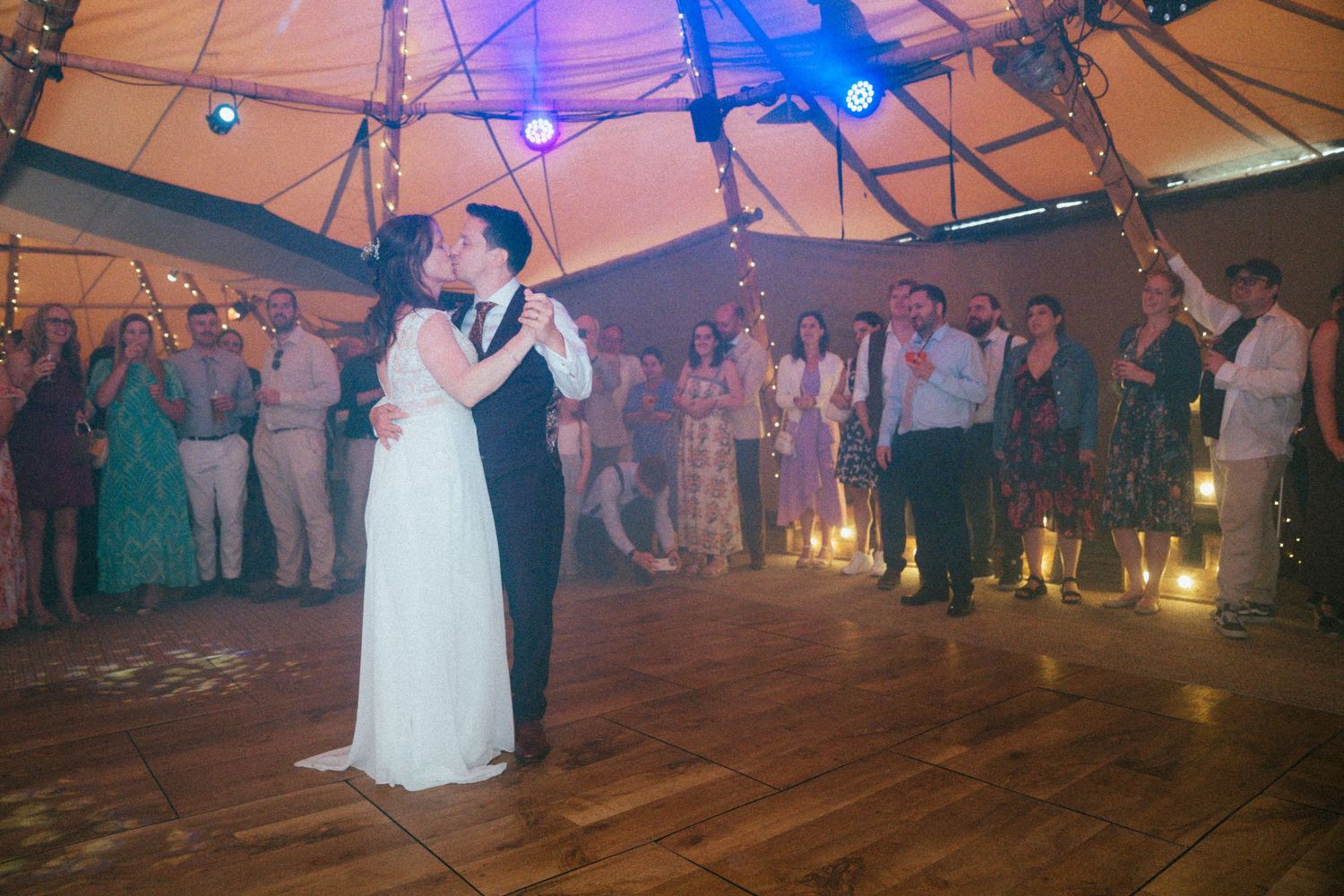Couple shares first dance under tent with warm lights as wedding guests watch and celebrate.