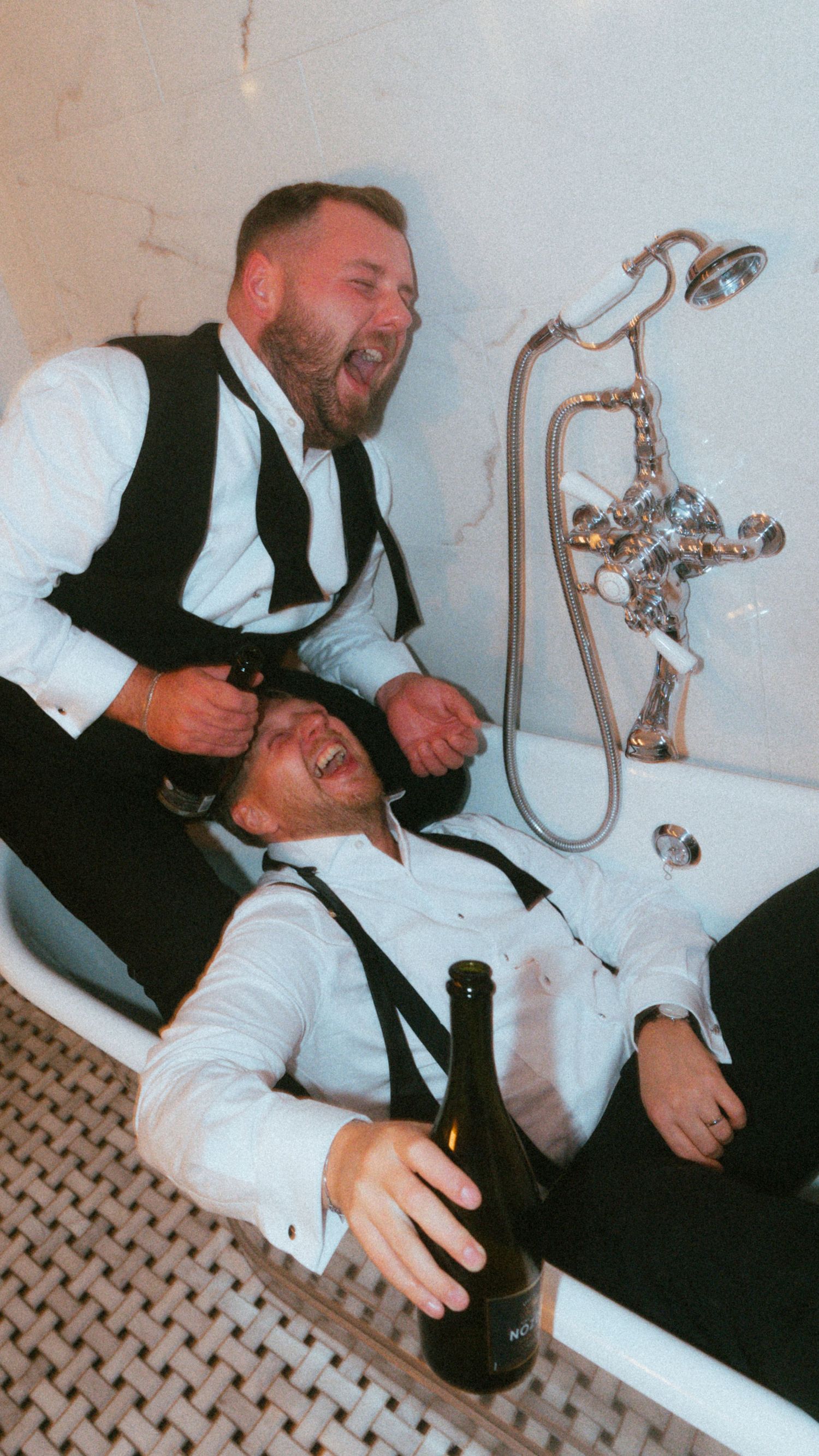 Two laughing men in vests and white shirts play in a vintage clawfoot bathtub with a champagne bottle, one pouring a drink onto the other.