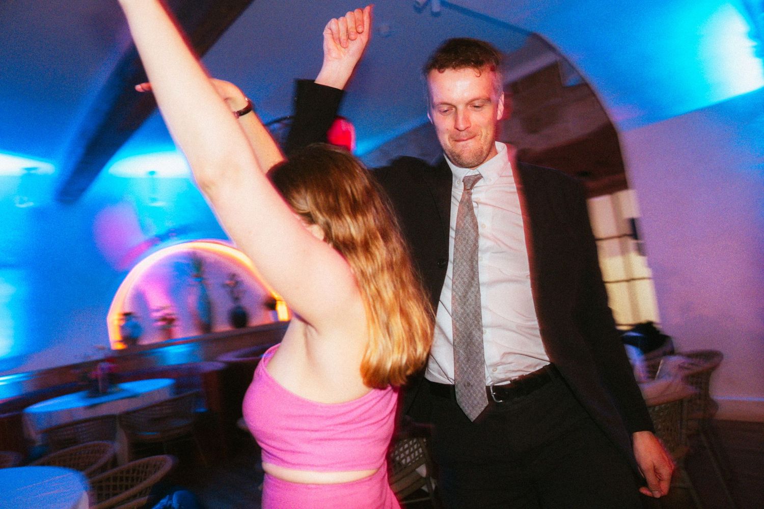 Woman in a pink top raises her arms while dancing with a man in a suit and tie at a lively wedding reception.