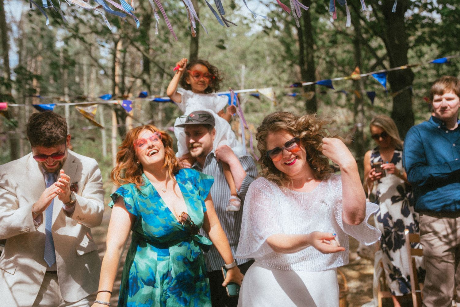 Joyful wedding guests dancing and celebrating outdoors in a forest venue decorated with colorful bunting flags.