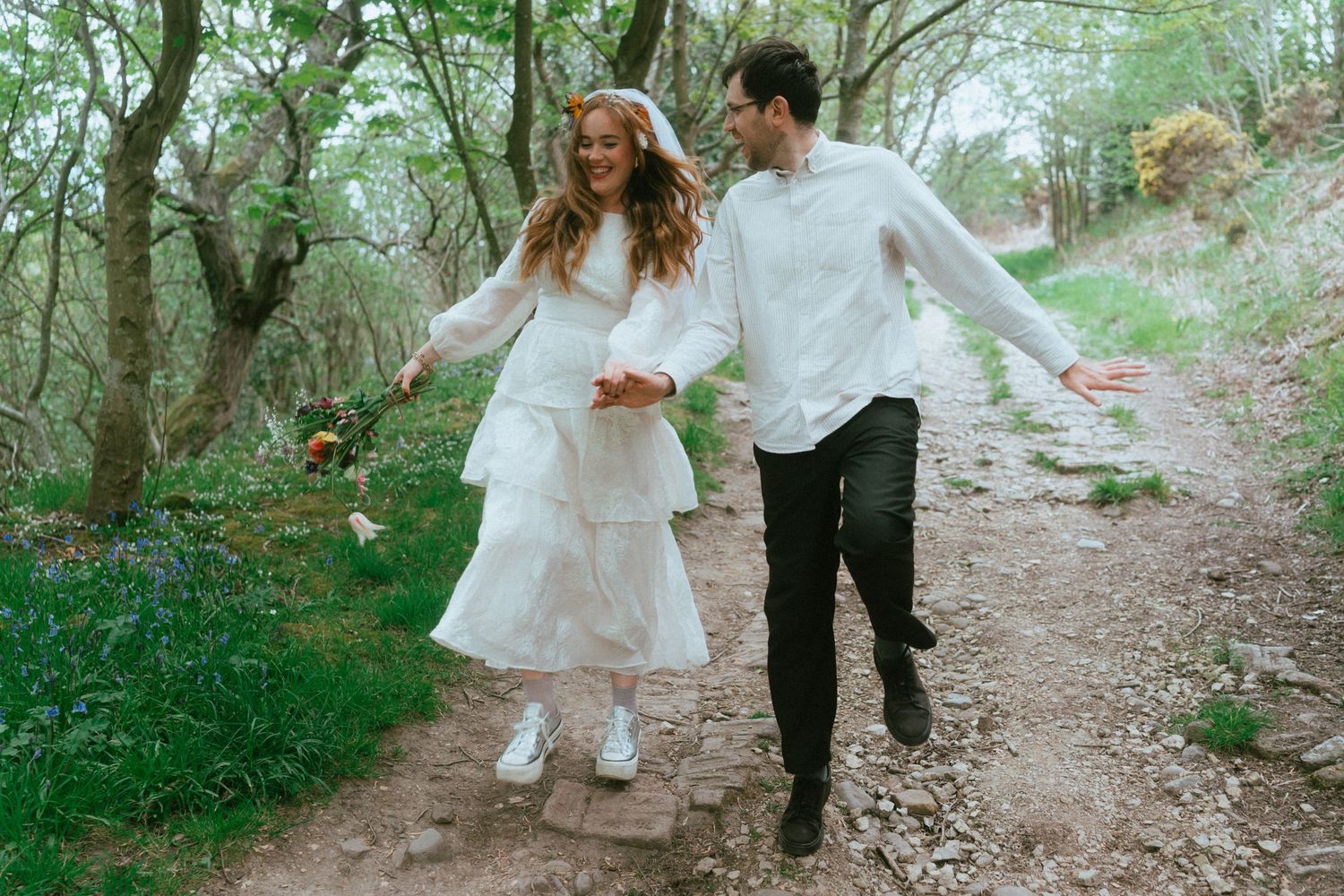 A smiling couple in white outfits holds hands and walks joyfully along a rocky woodland path surrounded by trees and bluebells.