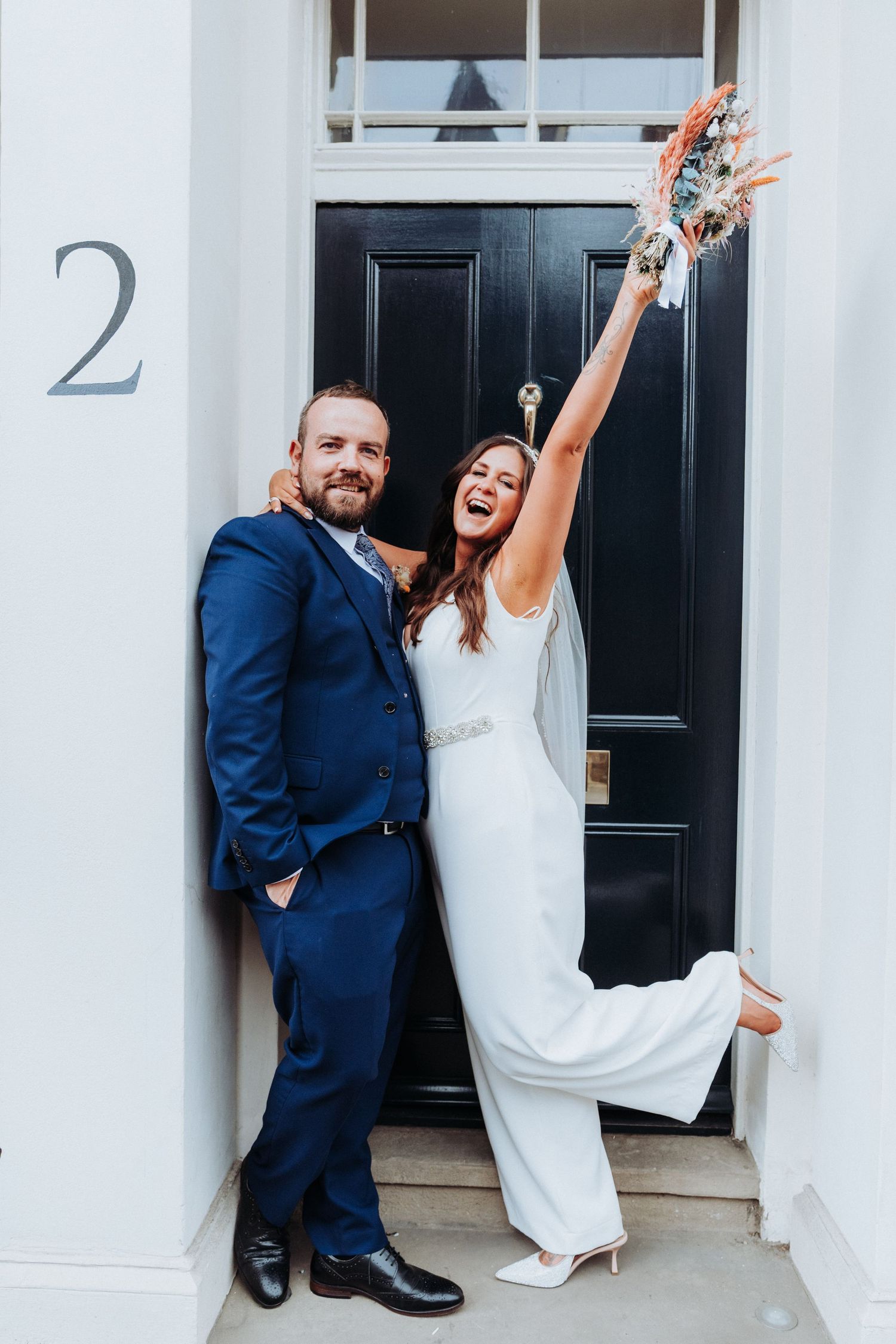 Joyful bride in white gown raises bouquet beside groom in navy suit at elegant black door numbered 2.