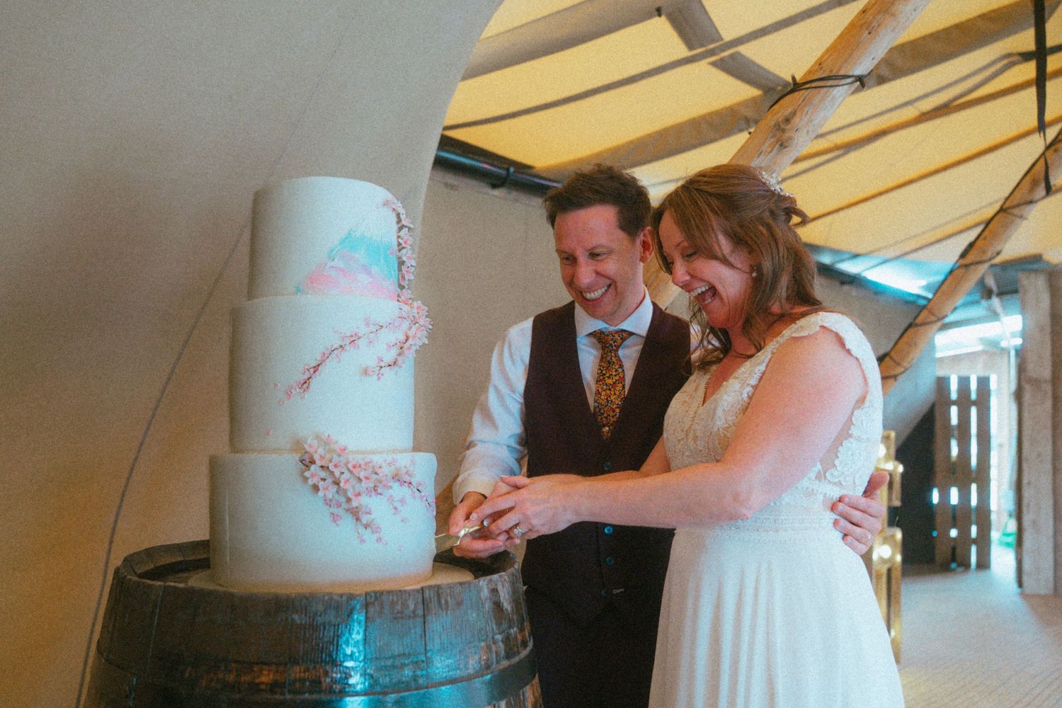 Bride and groom cutting a elegant three-tier white wedding cake decorated with pink floral details on a barrel.