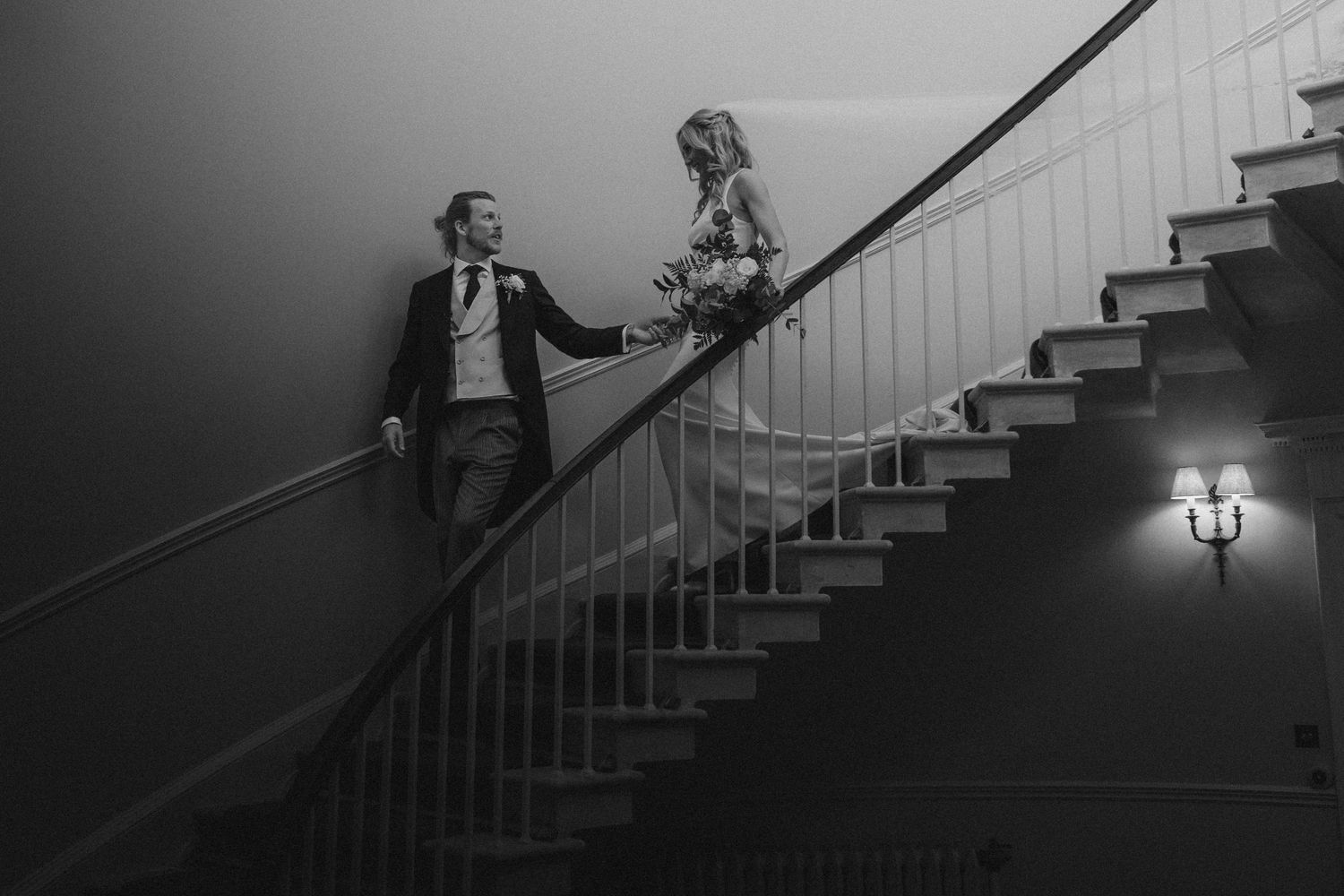Bride and groom hold hands on an elegant staircase in a stunning black and white wedding photo.