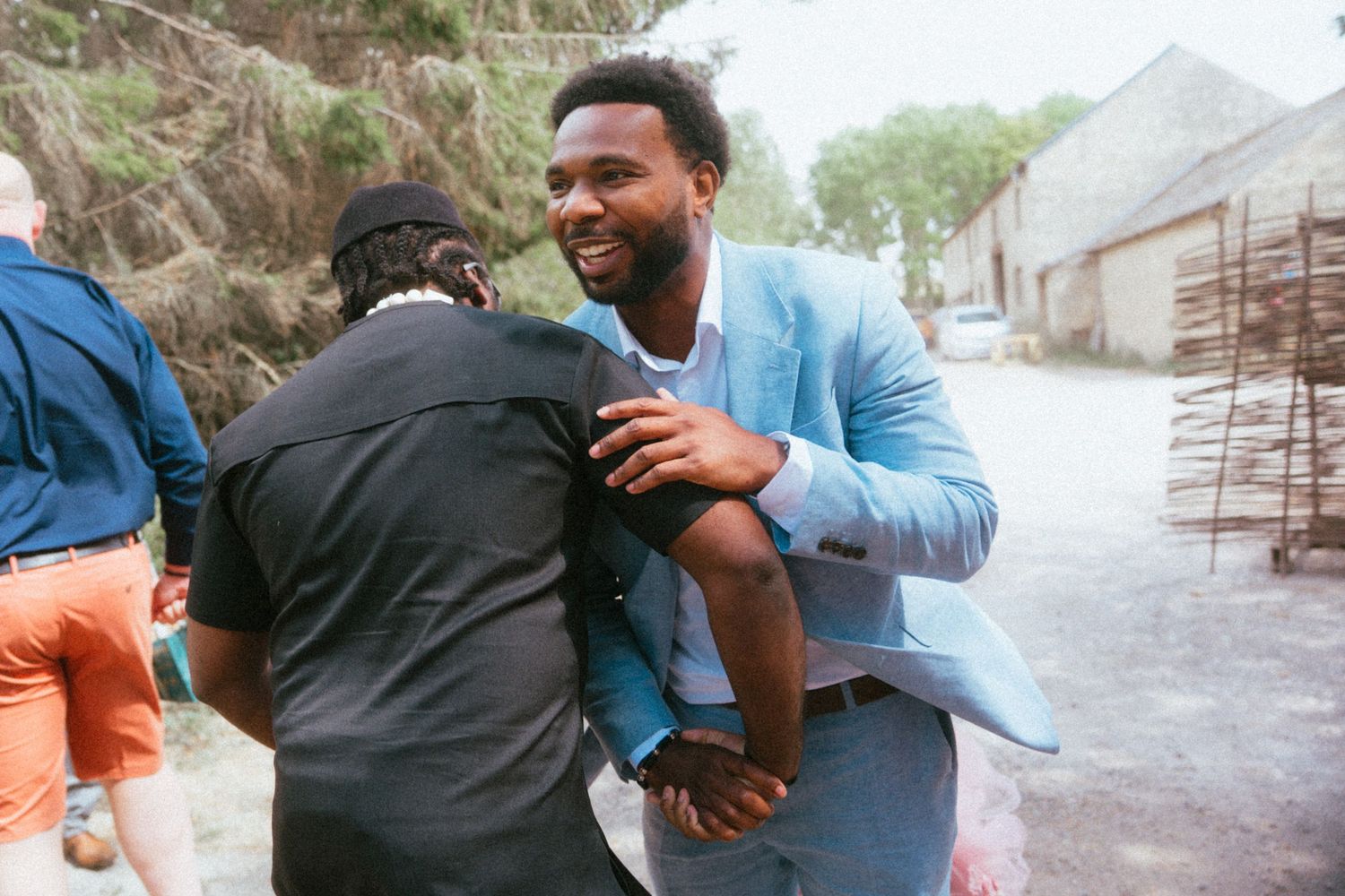 Two men in smart casual attire share a warm embrace outdoors at a wedding celebration.