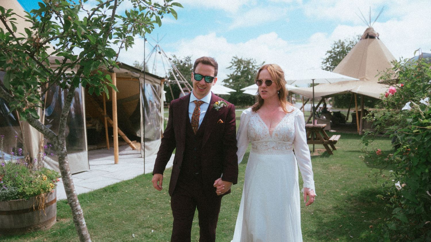 Newlywed couple walks hand in hand through garden with tipi wedding tent visible in background.