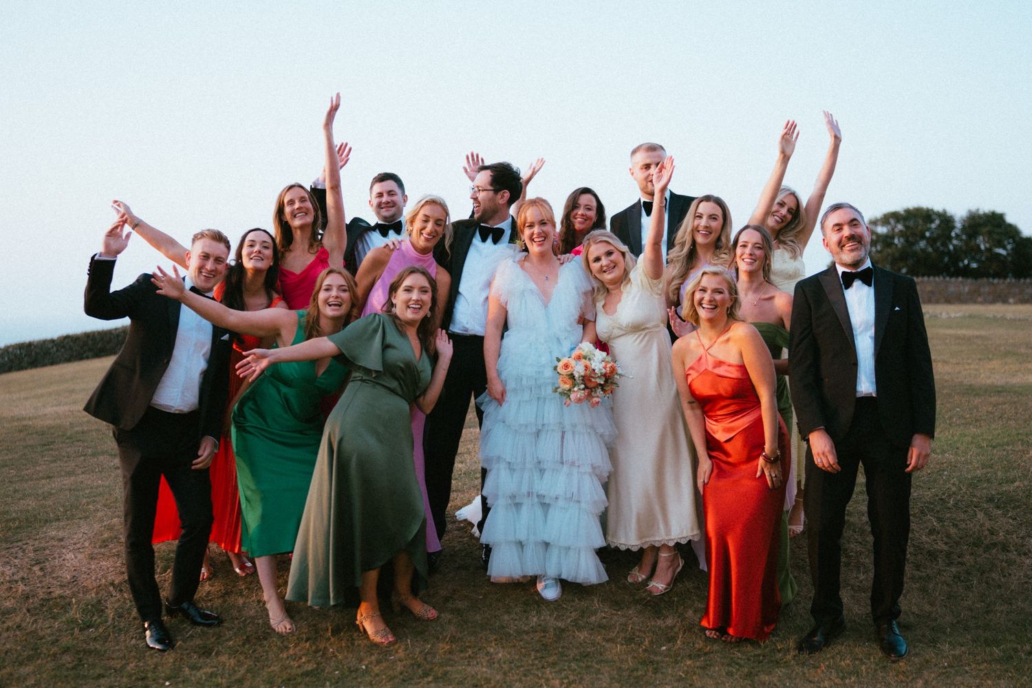 Wedding party group photo with the bride in a blue ruffled gown surrounded by cheering guests in formal attire at sunset.