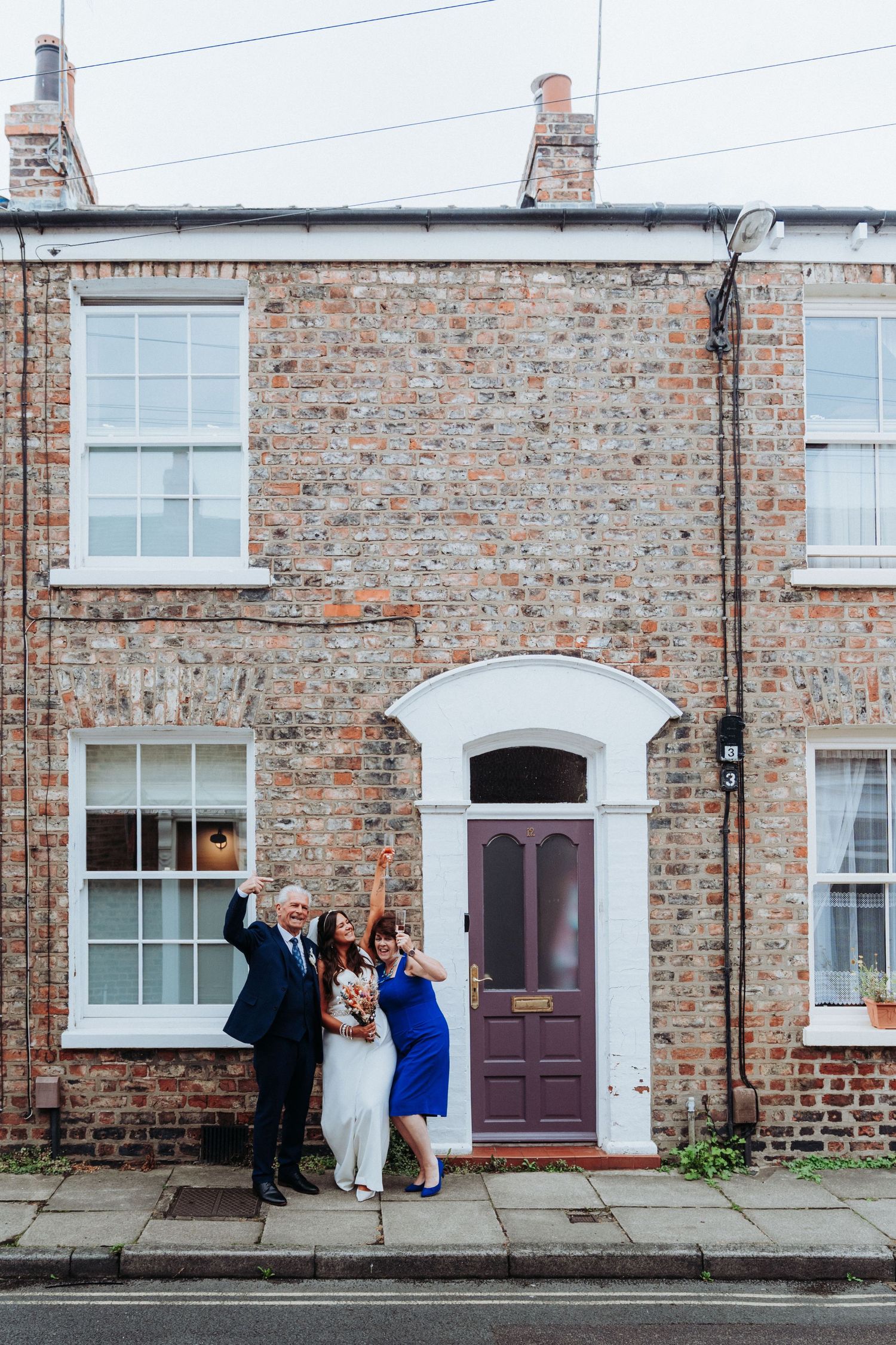 Bride in white jumpsuit celebrating with family outside a brick house with purple door.