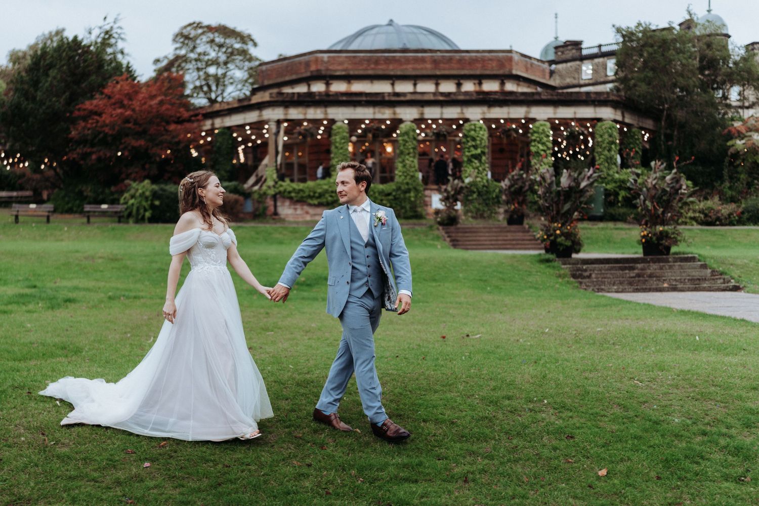 Bride and groom holding hands walking across lush garden lawn with fairy-lit venue in background.