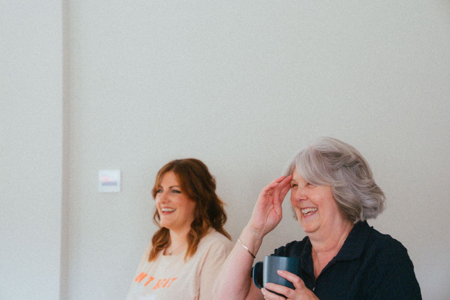 Two women laughing and smiling together, one holding a mug, in a bright modern room.