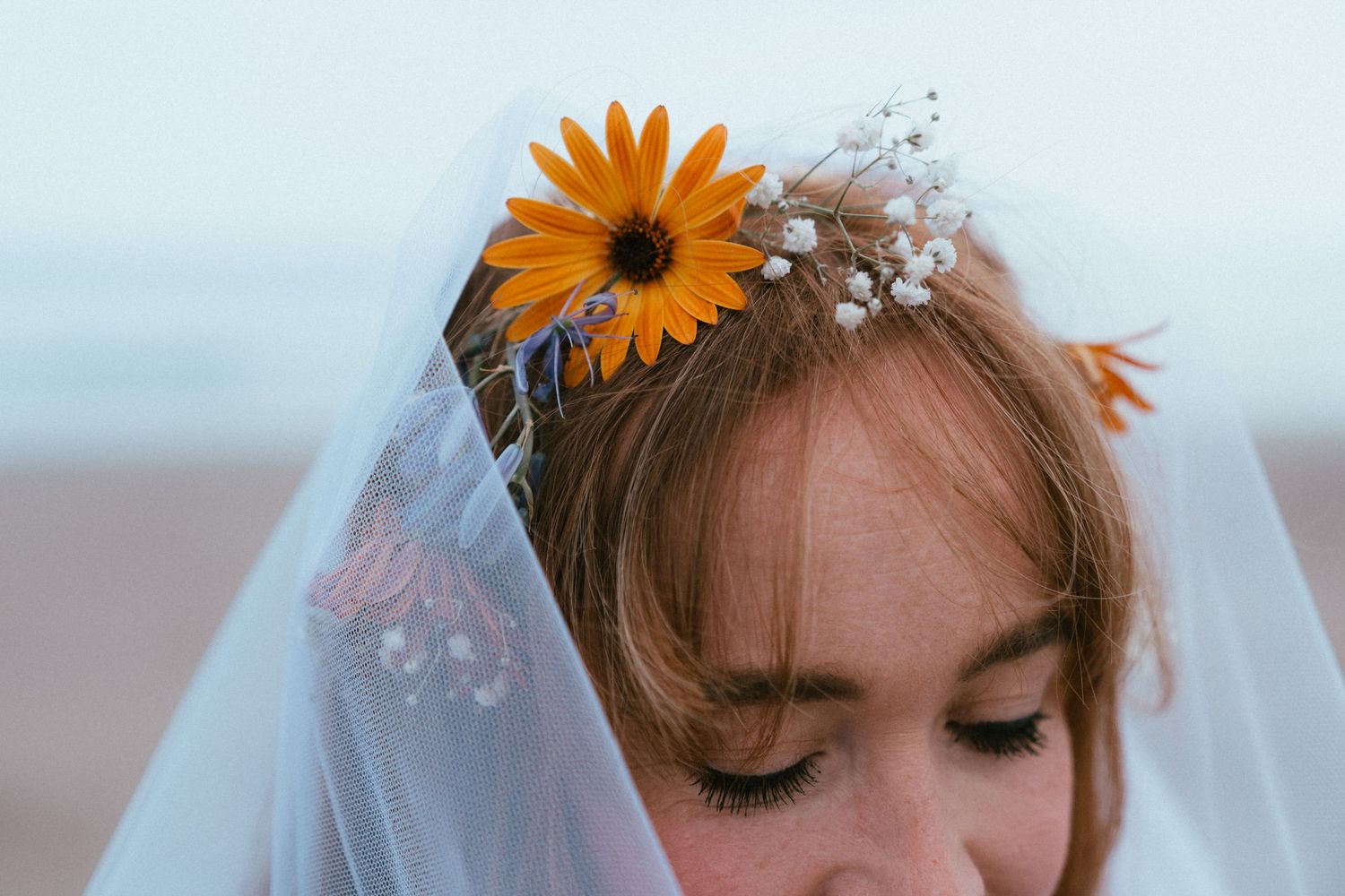 Close-up of a red-haired bride with downcast eyes wearing a sheer veil and a floral crown of orange daisies and baby's breath on a beach.