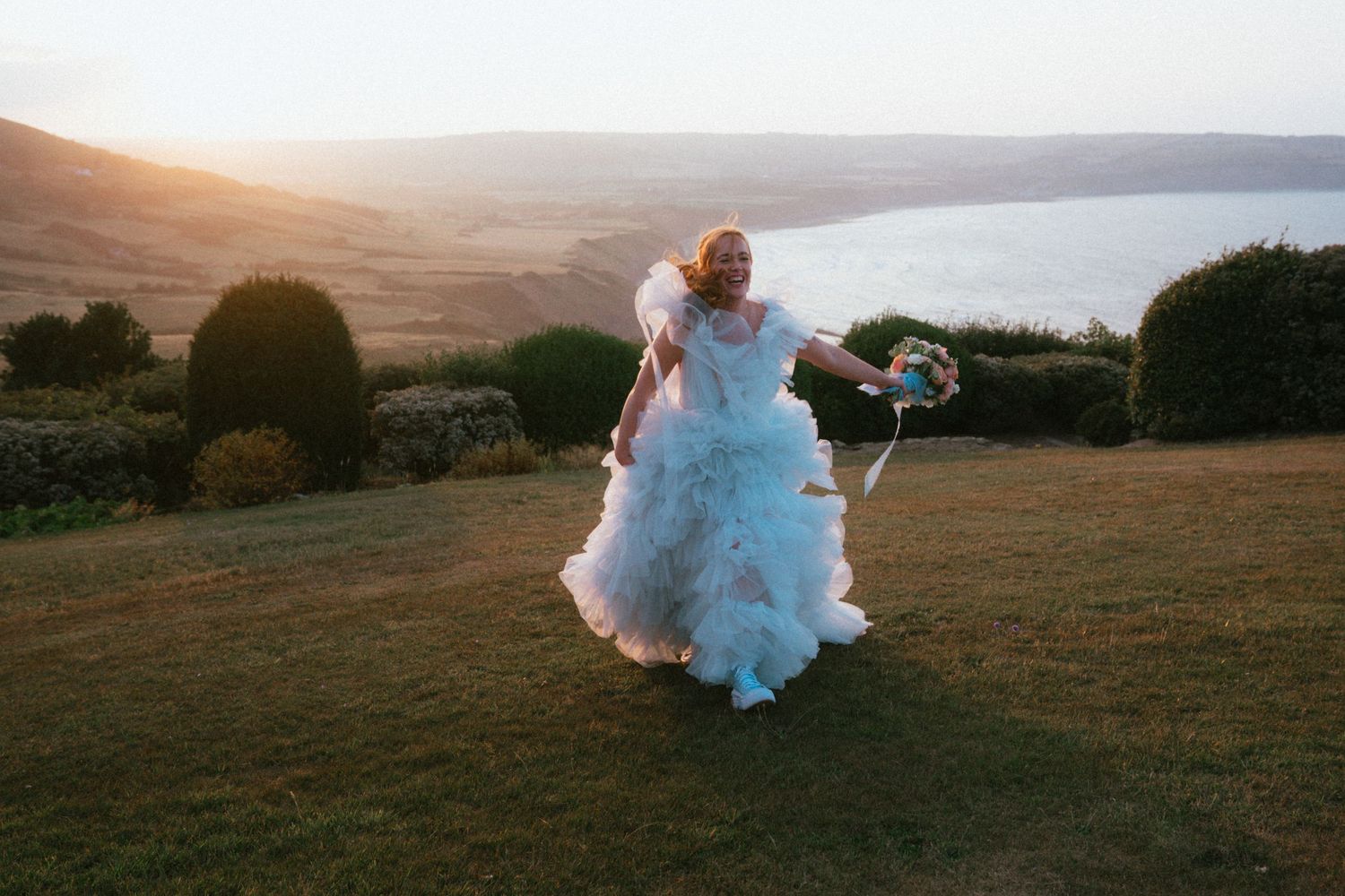 Bride in a flowing tulle wedding gown holds a bouquet while twirling on a grassy hillside at sunset with coastal views.