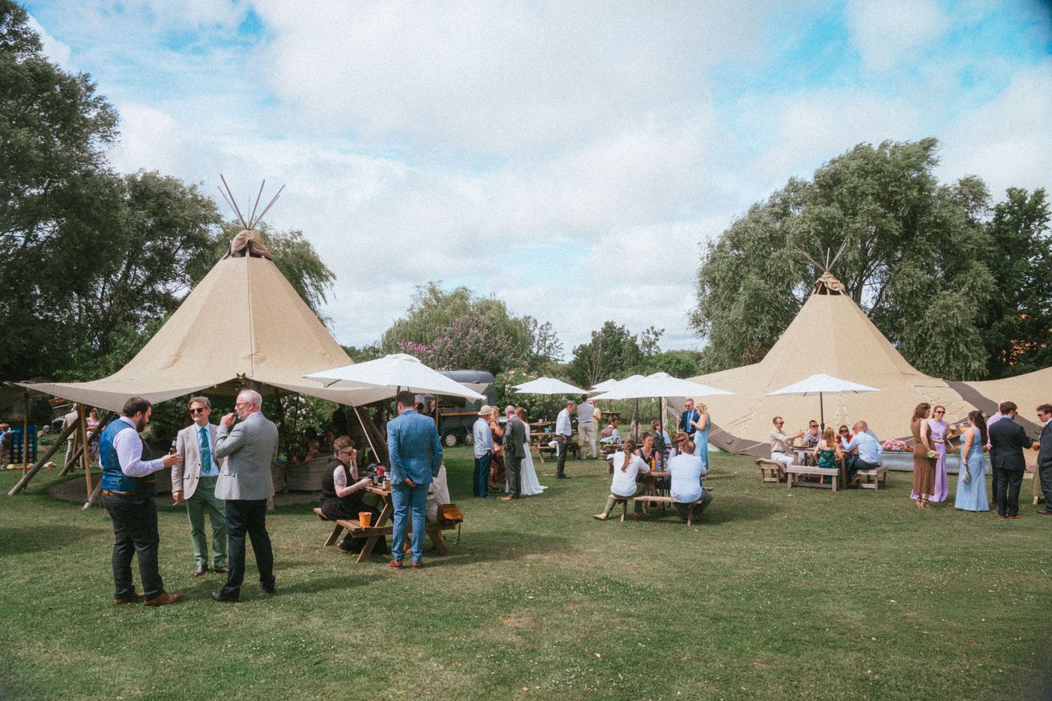 Wedding guests mingle on sunny lawn beside large Nordic tipi tents at outdoor summer reception.