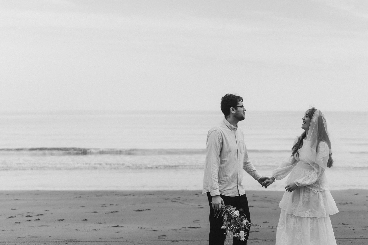 Black and white photo of a couple holding hands on a beach, dressed in wedding attire with ocean waves behind them.