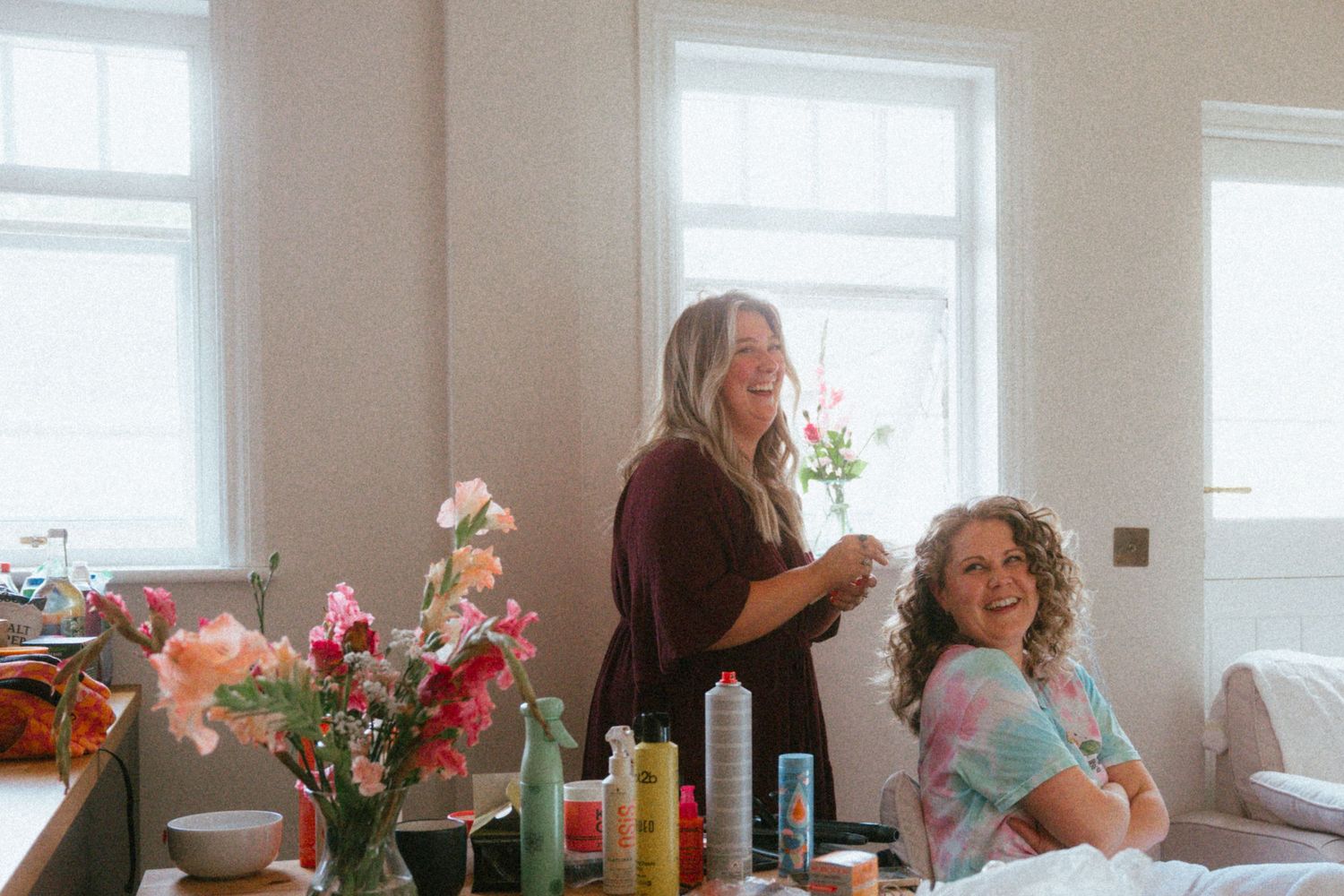 Two women smiling in a bright room with floral arrangements, beauty products, and a bed in the background.