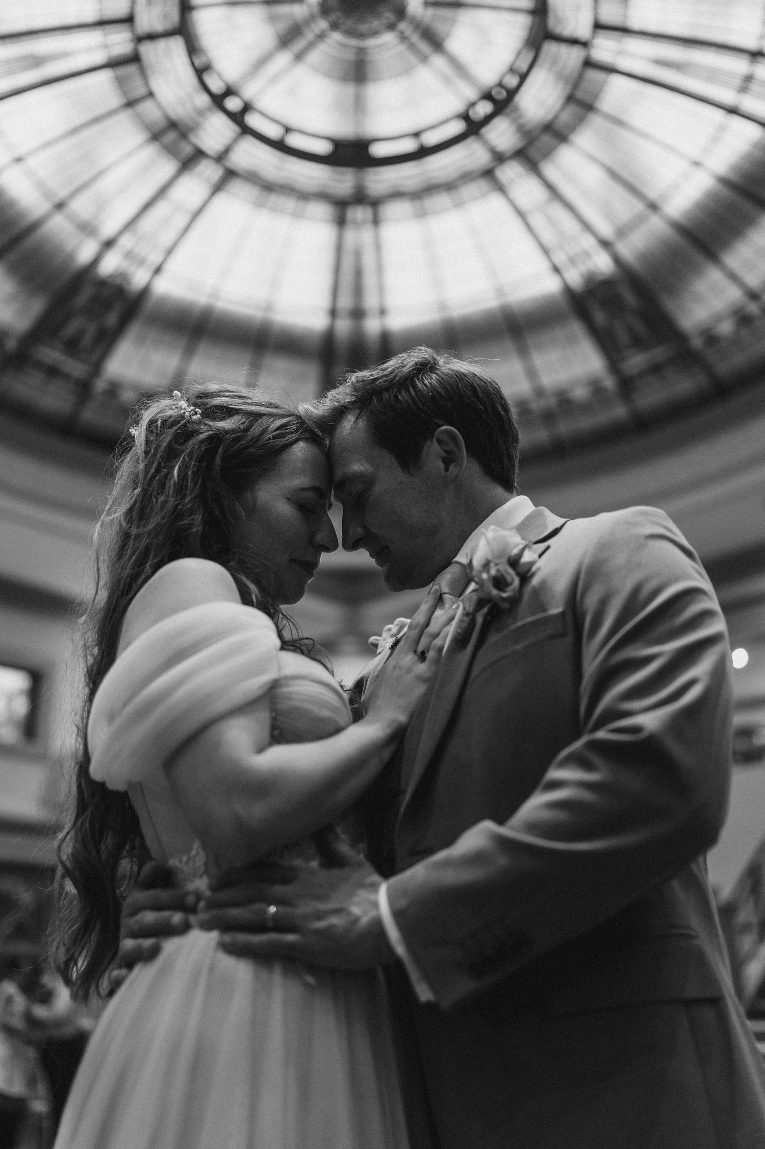 Black and white romantic photo of bride and groom sharing intimate moment under ornate glass dome.