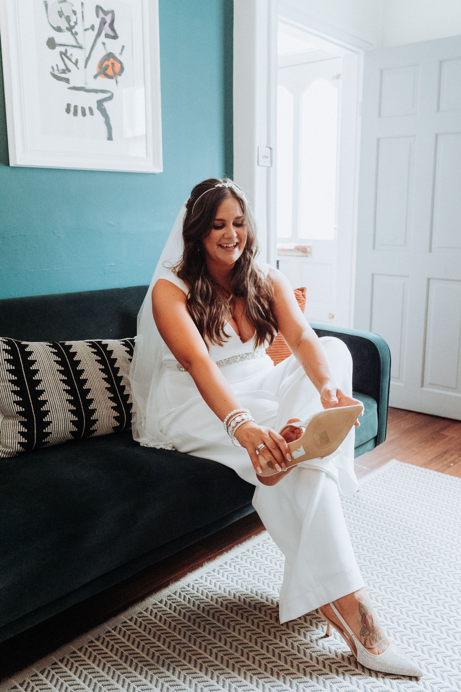 Smiling bride in white wedding jumpsuit sitting on dark sofa putting on heels.